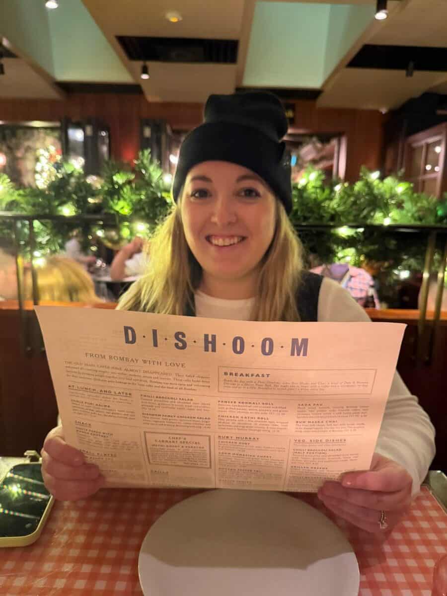 A woman holding a dishoom menu with greenery and ights in the background.