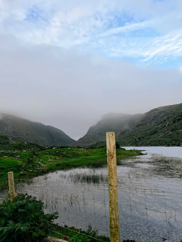 A wooden fence post overlooking a lake with foggy mountains in the background in the Gap of Dunloe. 