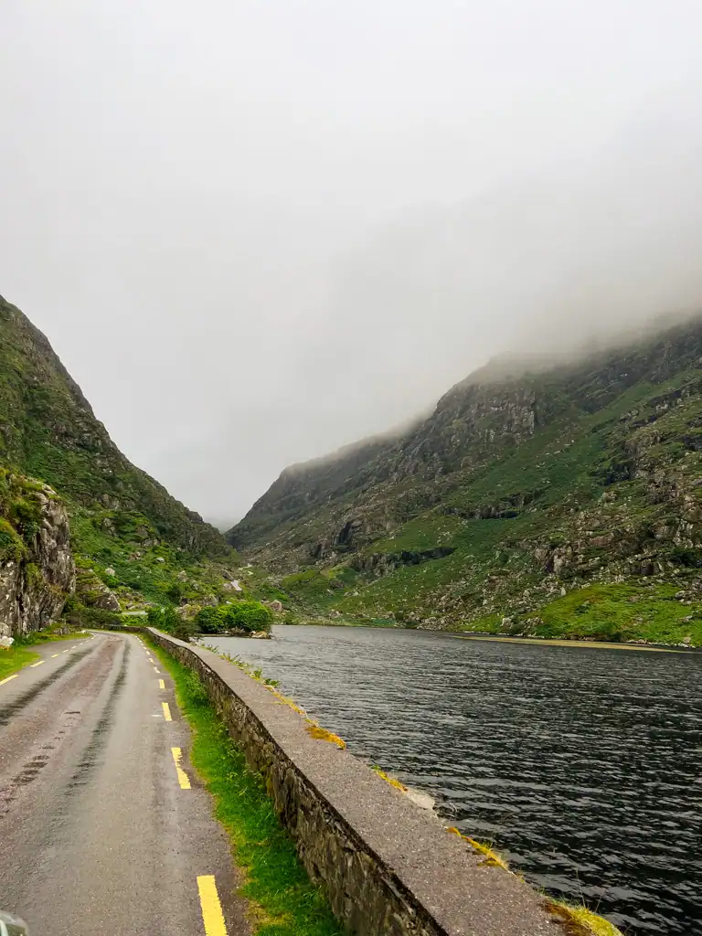 A narrow road next to a lake divided by a stone wall with foggy green mountains, in the Gap of Dunloe. 