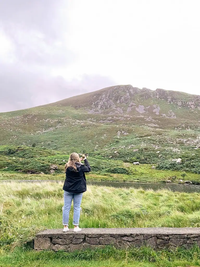 Woman stands on cement wall to capture a photo of the area and mountain in the distance.