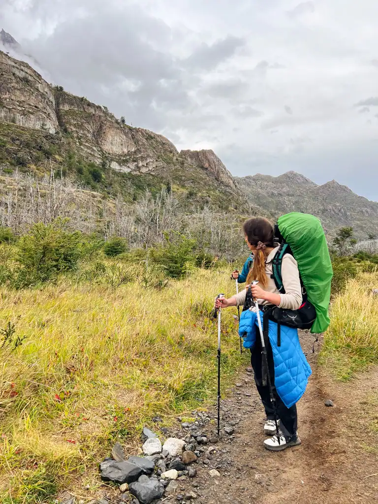 a girl stands with hiking poles, a blue coat and her backpack with green rain fly on top. 