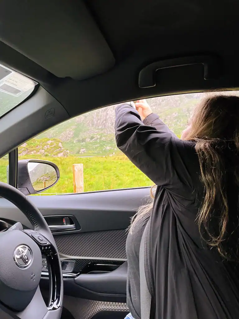 Woman takes photo out window of rental car while driving on the left in Ireland. 