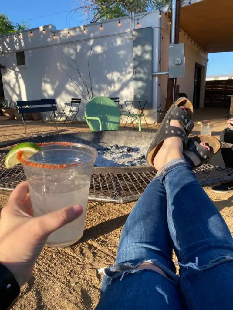 A hand holding a plastic cup with a clear liquid, tajin rim and lime. Her legs are laying out on a fire pit. 