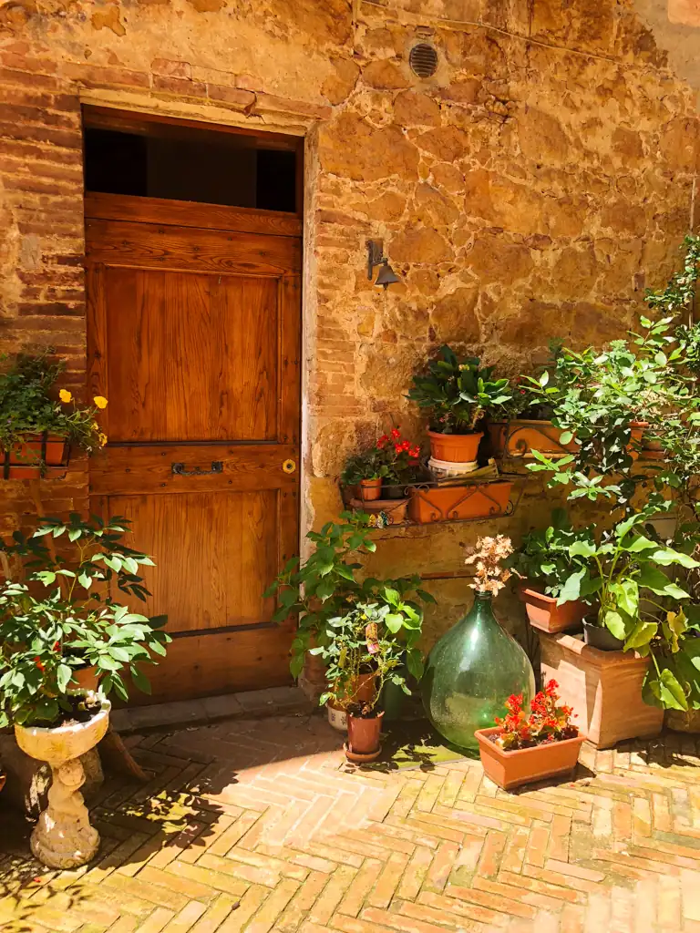 A doorway in Tuscany that is in a stone building and is surrounded by green plants and flowers in unique vases. 