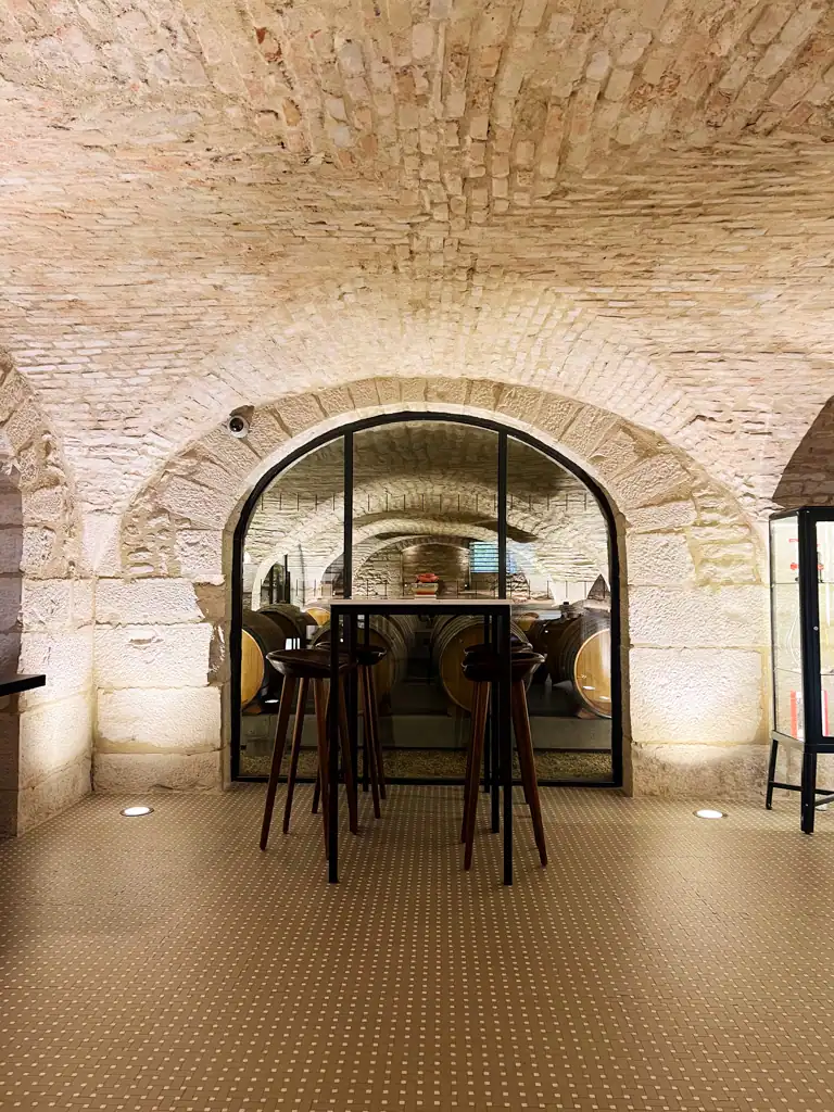 Table and chairs in a white brick wine cave with oak barrels through the window in the background. 