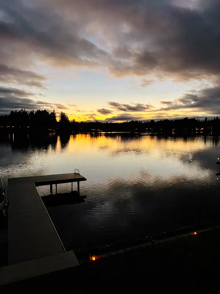 The sun almost all the way down with trees in the background, over a lake with a dock. 