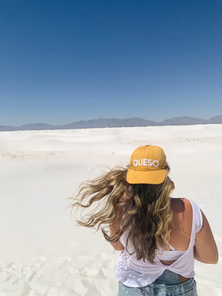 A white sand expanse at White Sands National Park on a National Park Roadtrip. A girl is in the forefront wearing a hat that says Queso.