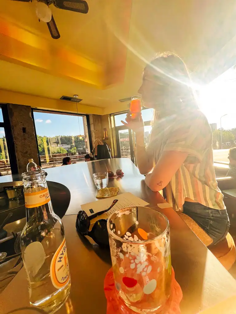 A girl drinks a cocktail, with another drink in the forefront and the sun shining in. 