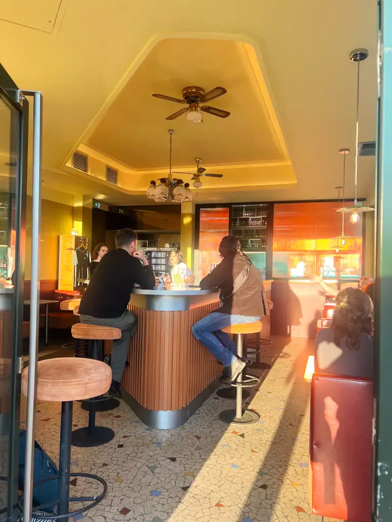 A narrow bar with the suns=light streaming in and people sitting on stools. 