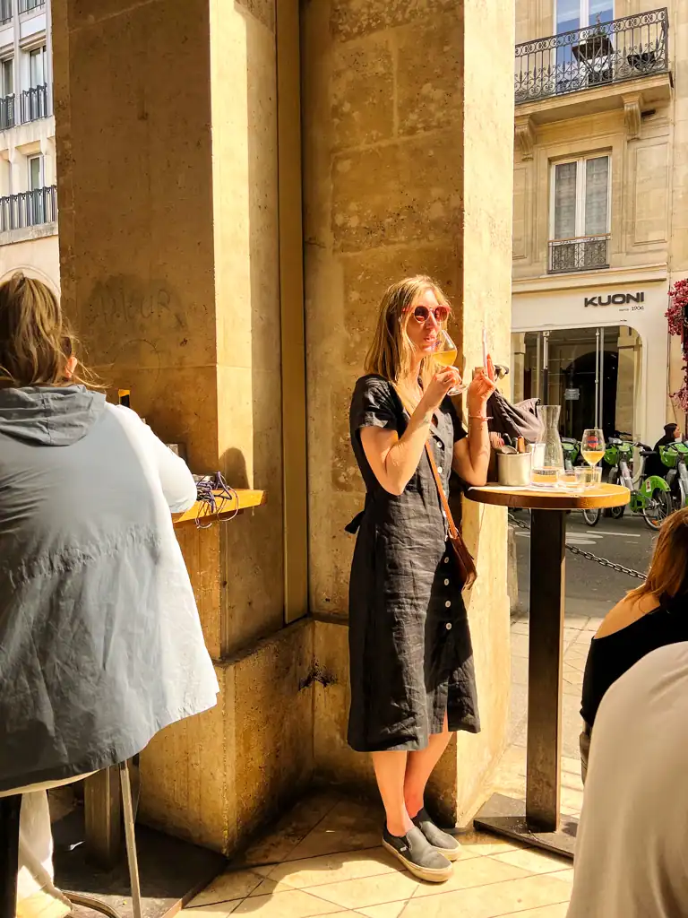 A girl in a black dress drinks wine while standing at a table. 