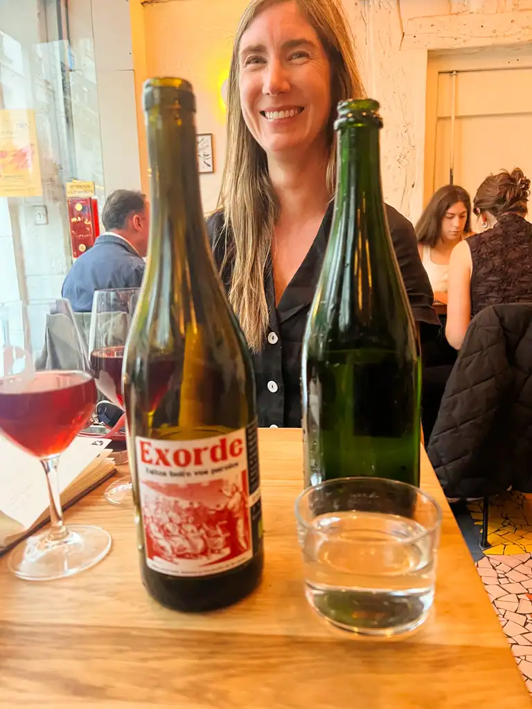 A girl is smiling and in the forefront is two bottles of wine. 