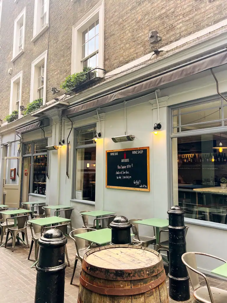 The outside of antidote wine bar with green topped tables, a chalkboard menu and wine barrel. 