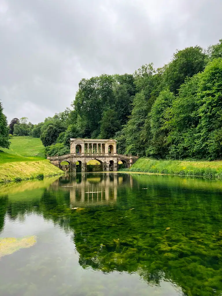 the palladian bridge in prior park in Bath seen on a weekend in bath The water is in the forefront and large trees and grass in the background.