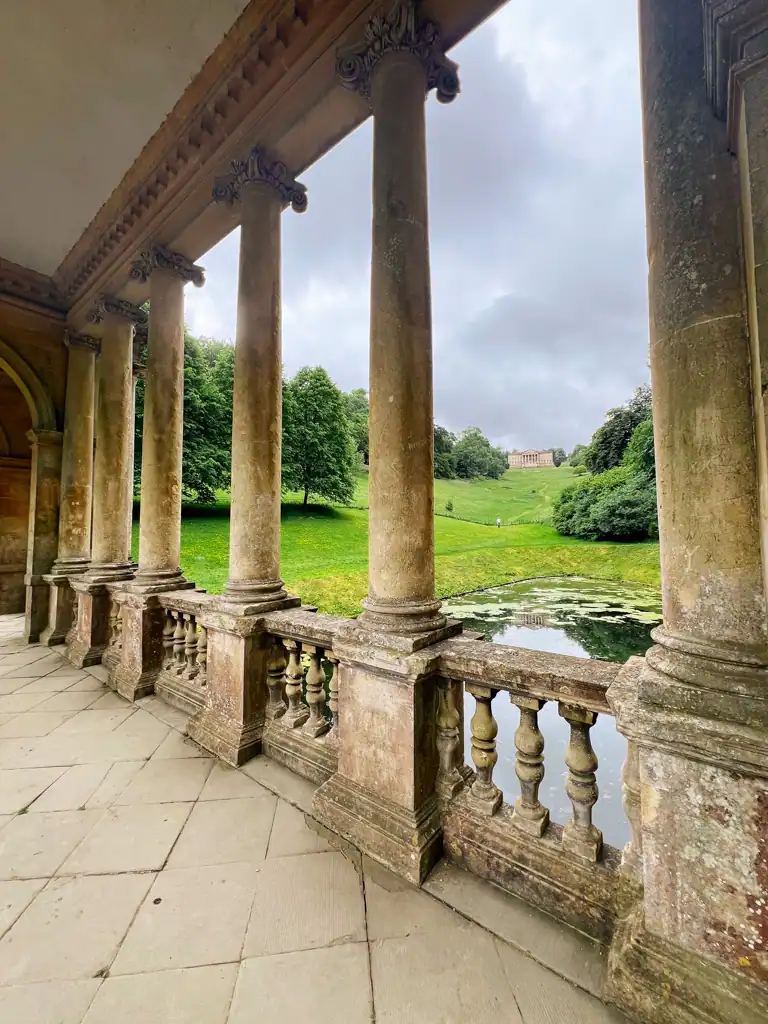 Standing on the palladian bridge in bath with ancient columns and stone with a lake and green hill in the background.