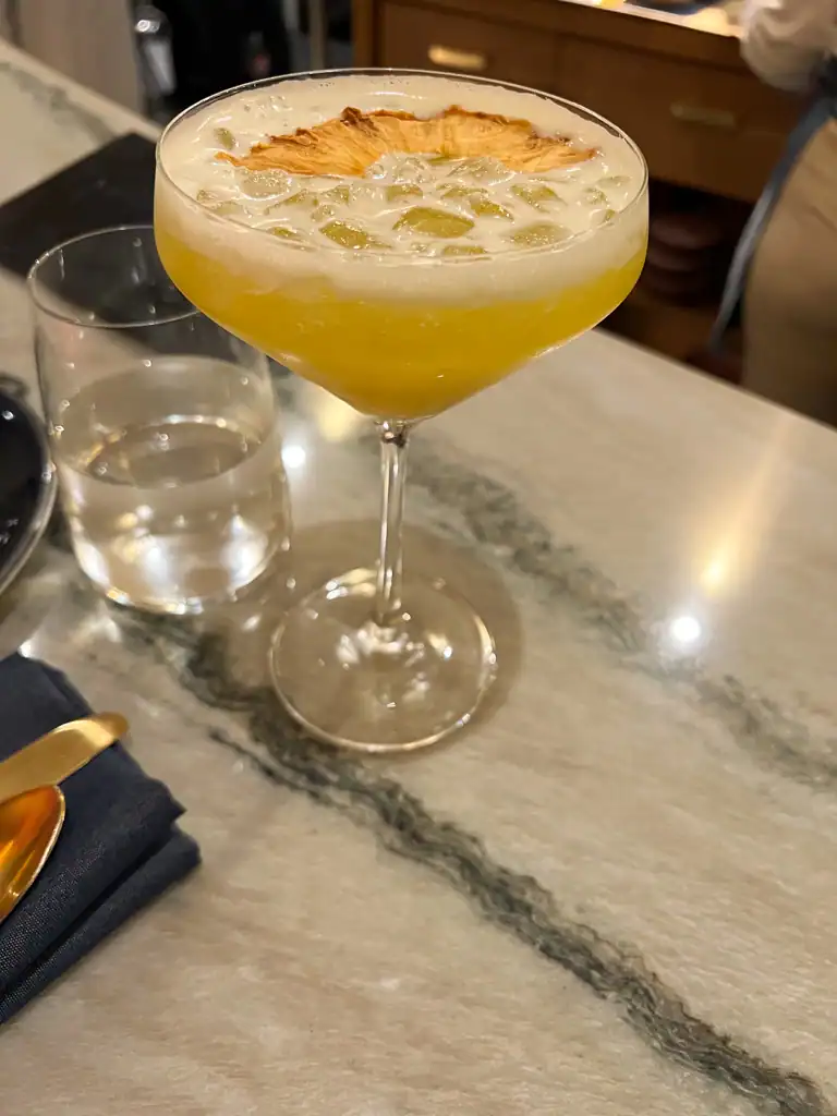 A yellow cocktail in a stemmed glass with a dried pineapple garnish. 