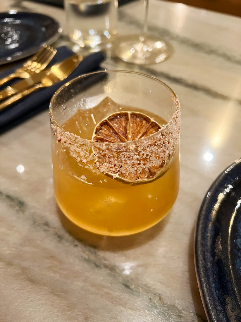 a yellow cocktail in a low ball glass with a dried lemon and a salted rim. 
