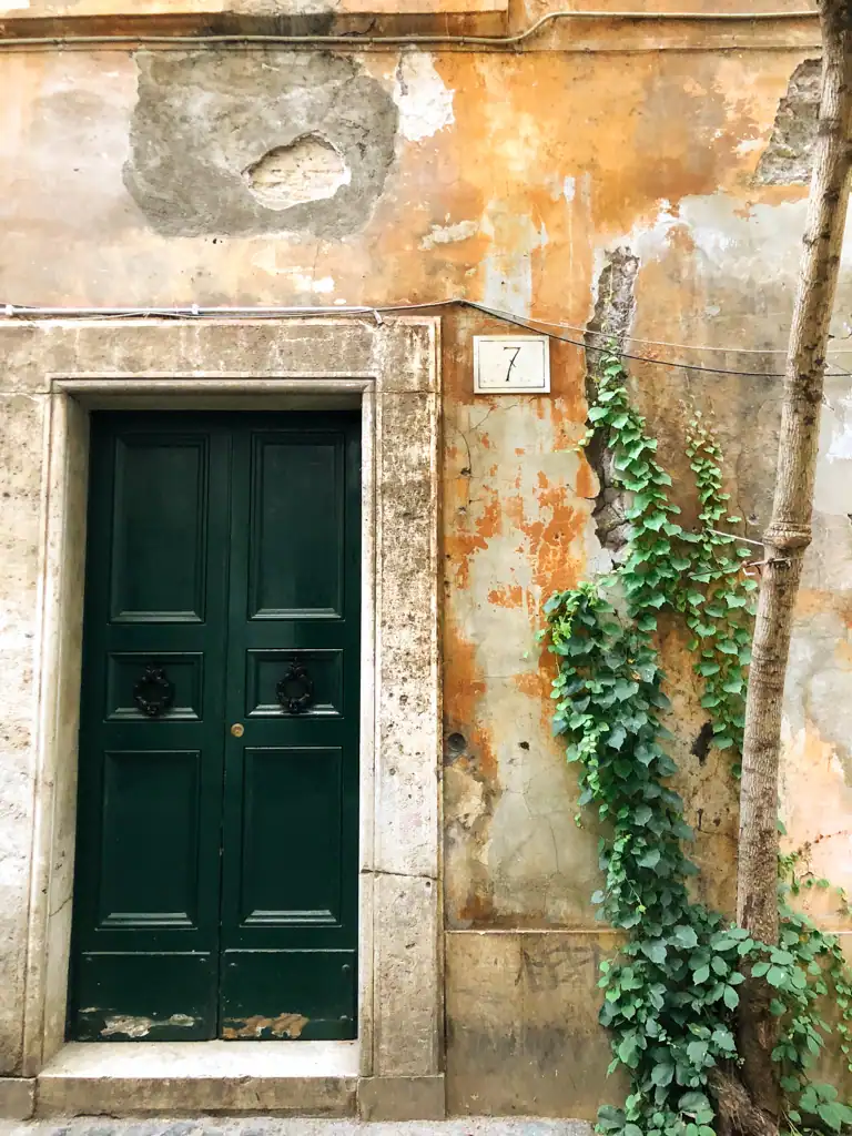 A dark green door with ivy climbing the wall of a wall in Rome that is rusty and falling apart. 