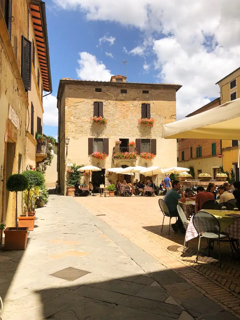 A small square with Tuscan buildlings and a two cafes with tables and umbrellas. 