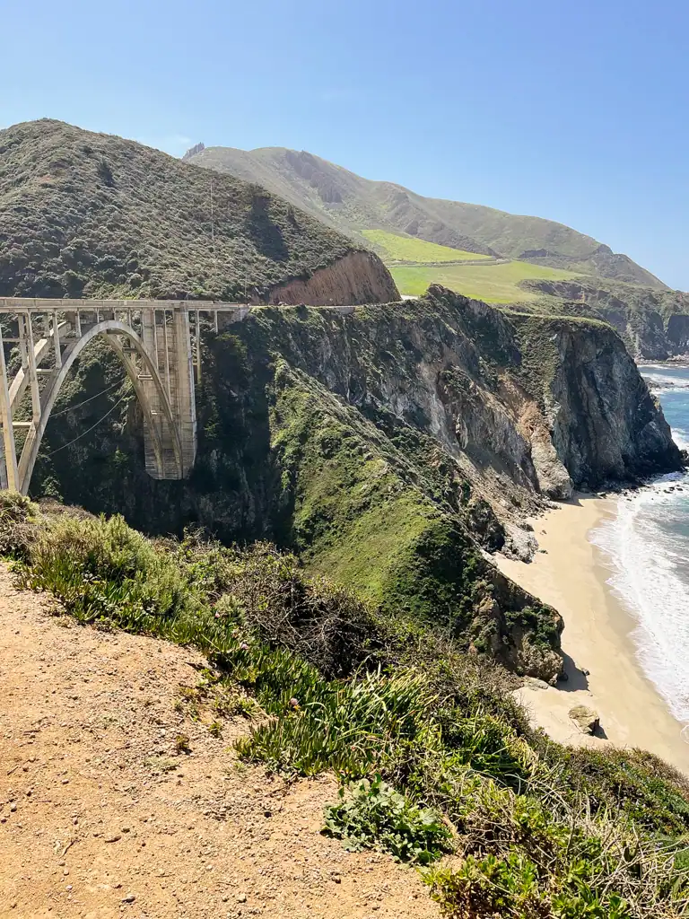 a photo of bixby bridge with green mountains in the back and the ocean peaking on the right. 