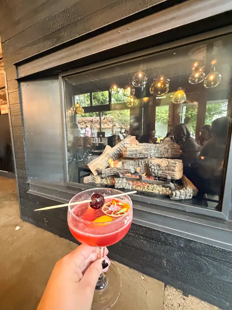A pink cocktail with a cherry on a stick in front of a fireplace. 