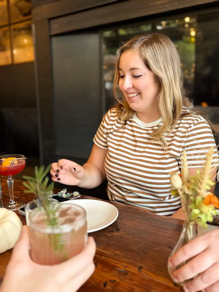 A girl in a white and tan striped shirt reaches out for her cocktail with a small vase and hand holding a drink in the foreground. 