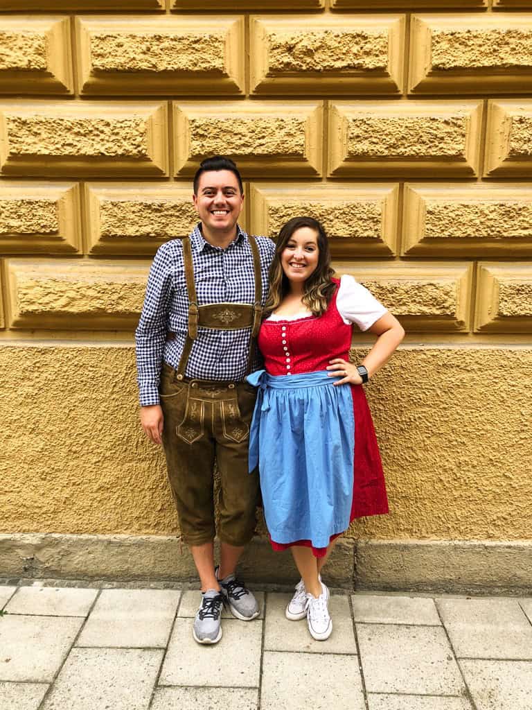 A man and women pose in traditional oktoberfest clothing against a brown brick wall. 