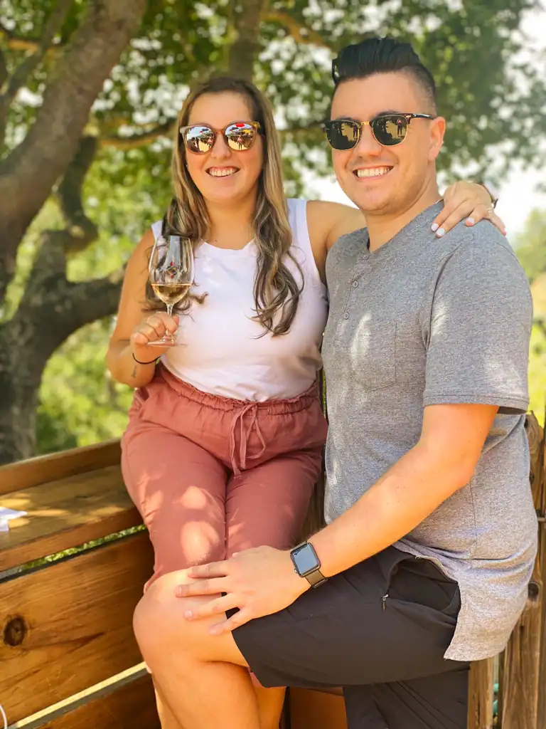 A couple poses with the woman holding a glass of wine both are wearing sunglasses. 