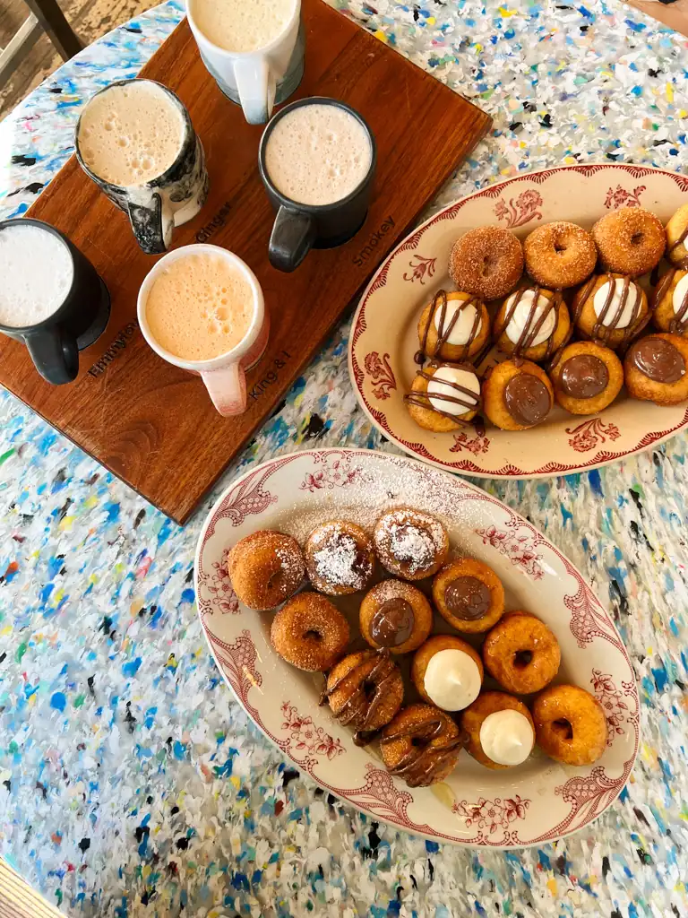 a wooden board with 5 cups of frothy chai on it and two plates of mini donuts with icings and chocolate. 