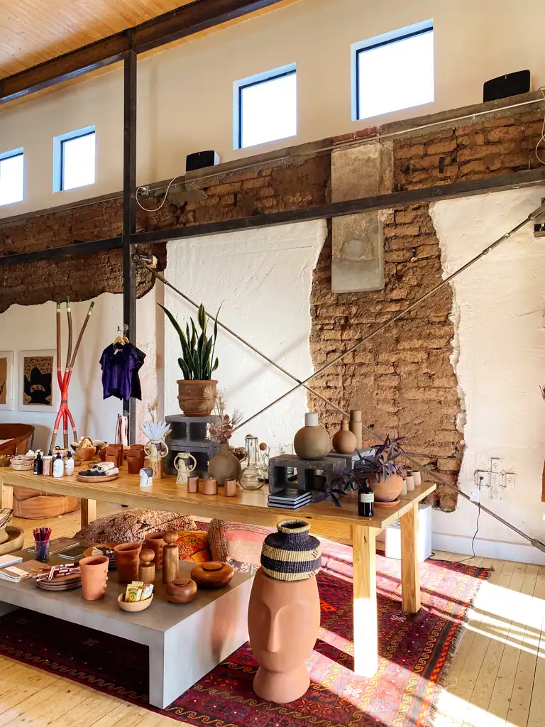 Pottery and plants lie on a table with a red rug beneath it and an exposed brick wall in the background. 