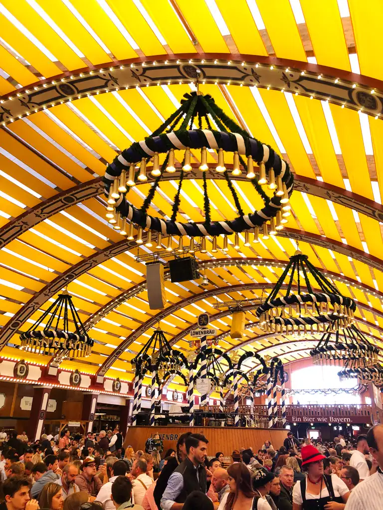 Inside of an oktoberfest tent with yellow slats on the ceiling and eccentric light fixtures. The tent is full of people.