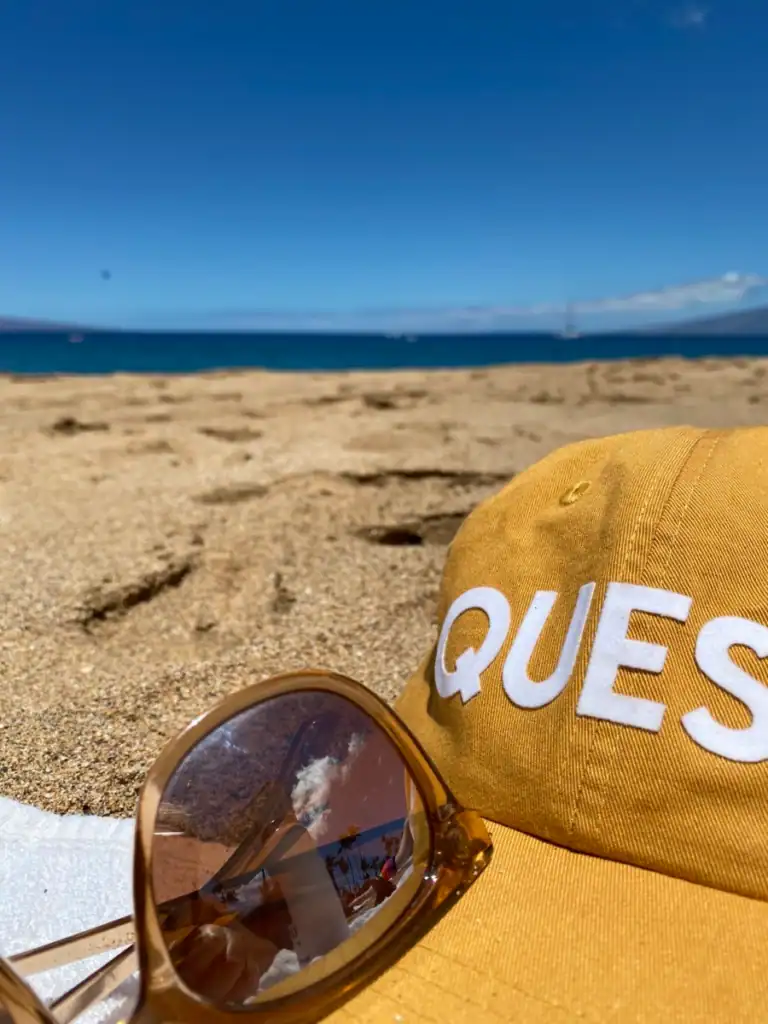 A yellow hat that reads "queso' next to sunglasses resting on sand near one of the 5 amazing beaches in Maui. 
