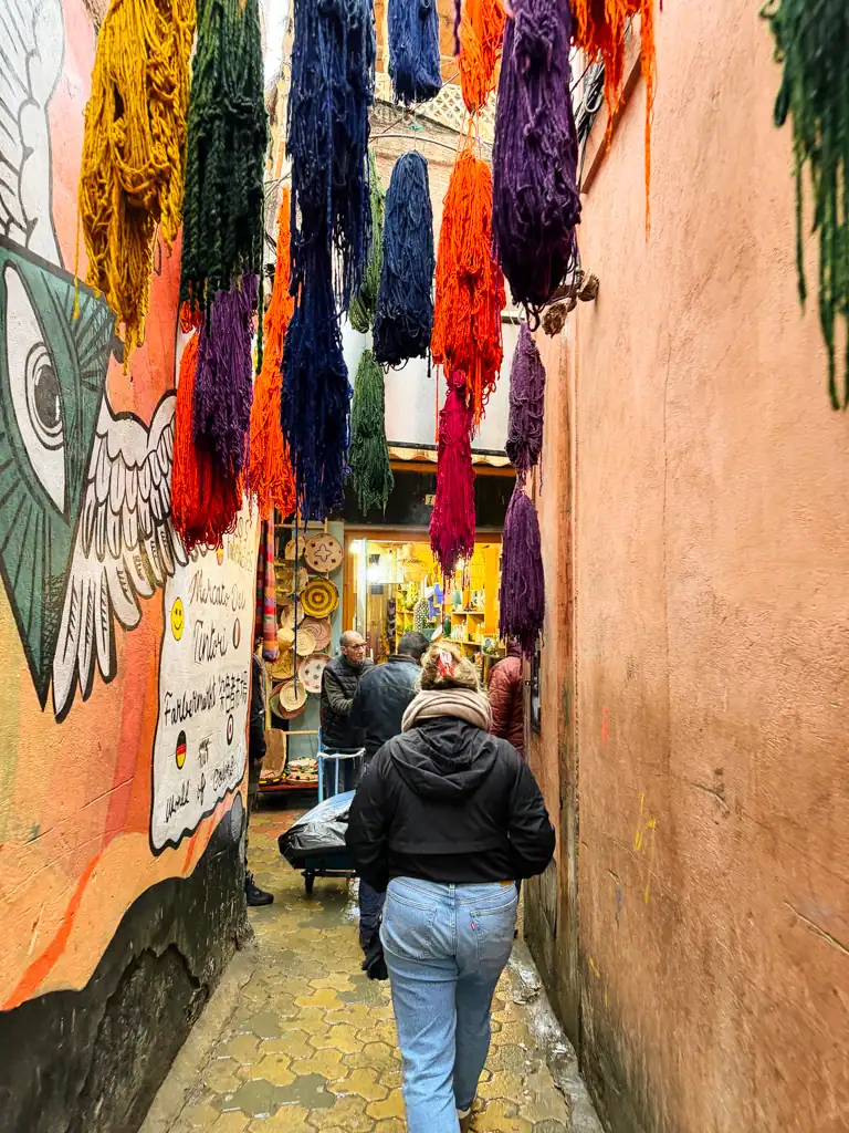 A girl in a black coat walks through the marrakech medina with colorful yarns hanging above her and a large mural with an eye and wing on the left. 