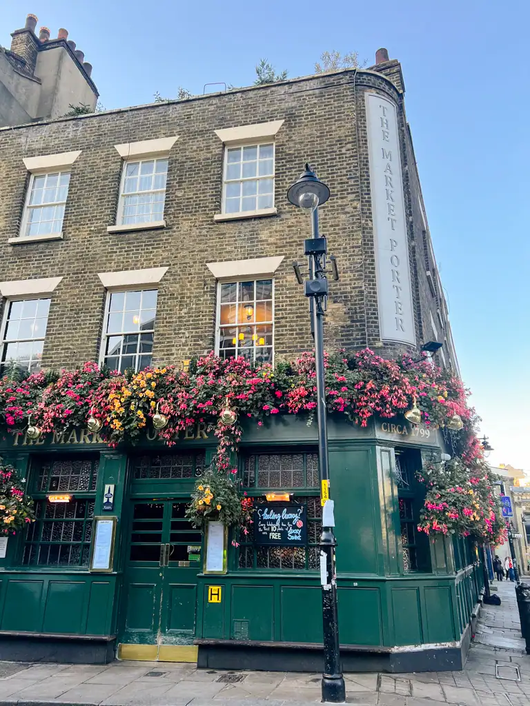 The outside of a green pub covered in flowers on a stone building, seen in a london guide. 