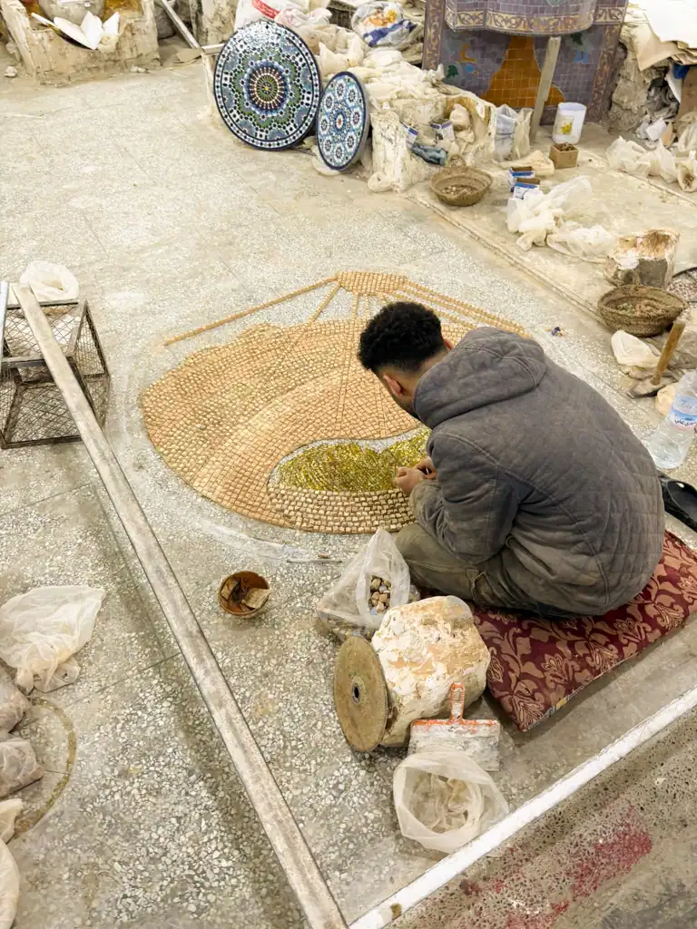 A man sits on the ground creating a mosaic placing each tile individually.
