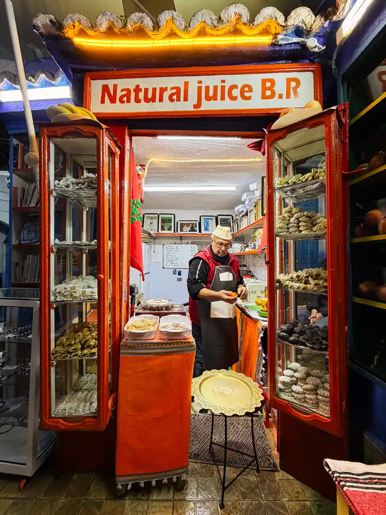A man in morocco makes fresh squeezed orange juice in his red shop with cookies in the display cases.