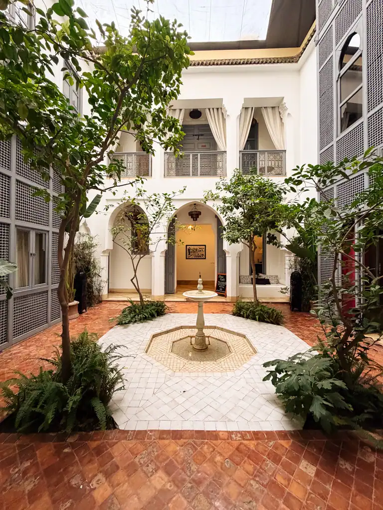 the center of a riad with white walls, archeways, white curtains and small fountain surrounded by trees.