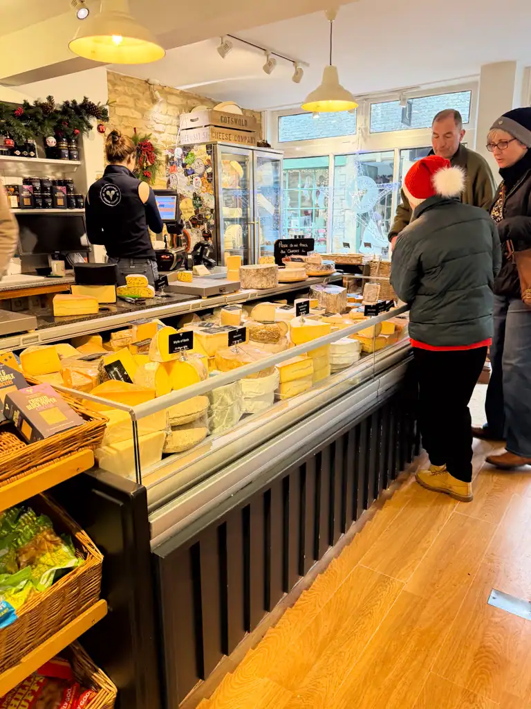 A cheese shop with a large display case of cheese with people standing in the corner. 