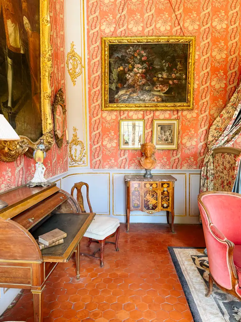 centuries old room with a antique desk, chair, red busy wallpaper and paintings on the wall. 