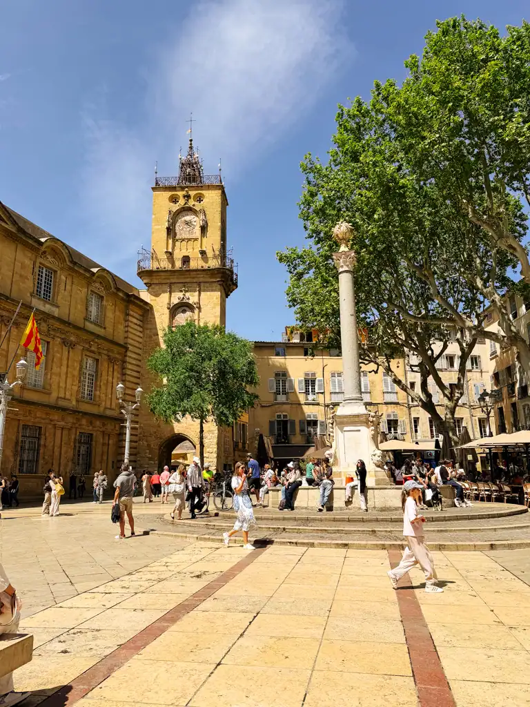 a sunny square with a fountain, people walking by and a clock tower in a weekend in aix-en-provence. 